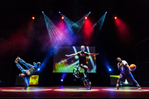 A group of athletic performers in various stunts using basketballs, bicycles and a giant hoop