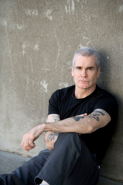 Photo Credit: Ross Halfin Henry Rollins in a black t-shirt, seated and leaning against a grey wall, photo credit to Ross Halfin
