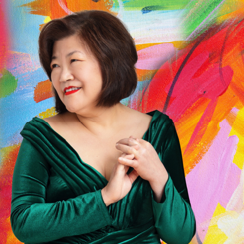 Alt text:  Person in a green velvet dress posing with clasped hands in front of a vibrant abstract painted background.