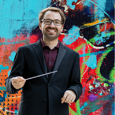 Alt text:  Person in a dark suit holding a conductor’s baton, standing before a colorful abstract art background.