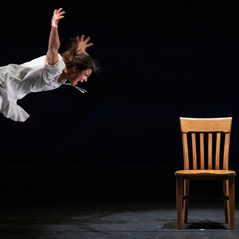 Dancer in white with arms up looking like she is falling on the left side of the screen with a empty wooden chair facing us
