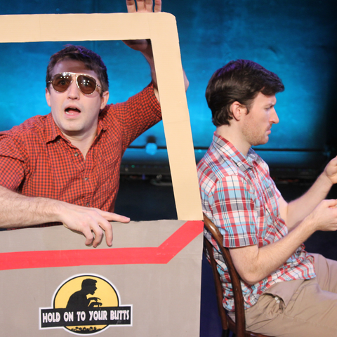 two performers in a 'jeep' with the one in the backseat wearing sunglasses and looking towards the camera