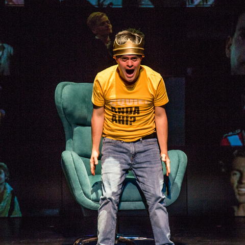 An actor dressed in a yellow short and jeans faces the camera with their arms at their sides