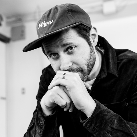 Dan Mangan in a ball cap, resting his lips on his hands in black and white