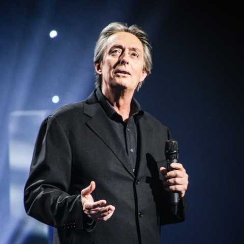 Performer in a dark suit holding a microphone on stage under blue lighting.