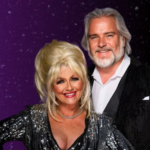 Two people smiling in front of a purple, starry background. The person on the left wears a sparkly black and silver outfit with an updo hairstyle, large earrings, and a necklace. The person on the right has light-colored hair and a beard, dressed in a white shirt and dark jacket.