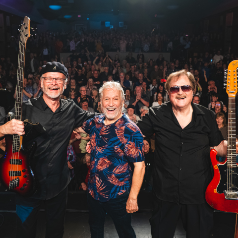 Three band members stand on stage holding guitars, smiling toward a large cheering audience in a dimly lit concert venue.