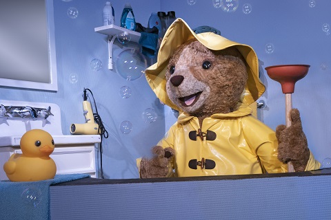 Paddington bear in the bathroom wearing a yellow slicker and carrying a plunger