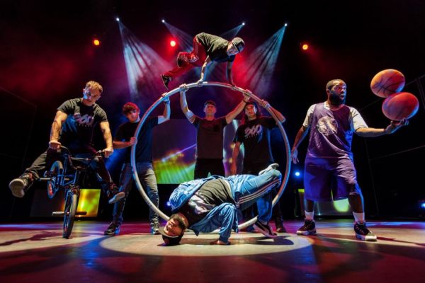 A group of athletic performers in various stunts using basketballs, bicycles and a giant hoop