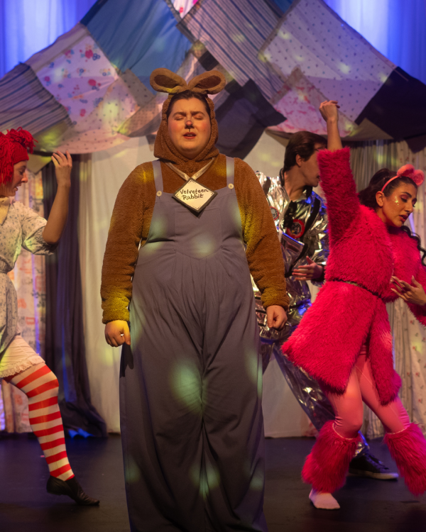 an actor in jean overalls looking at the camera with a women in pink running off towards the right