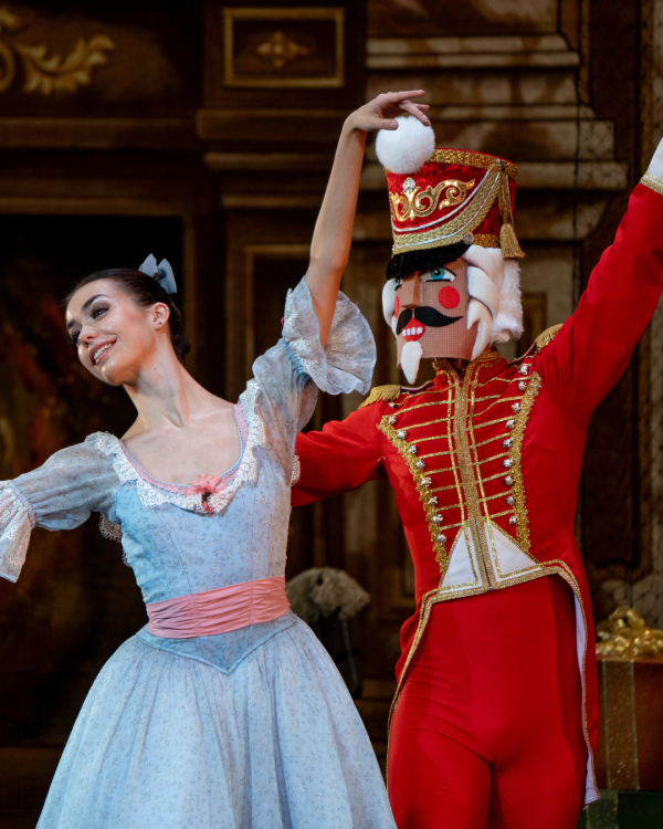 A dancer dressed as a nutcracker in red and another in a light blue dress pose with their arms up