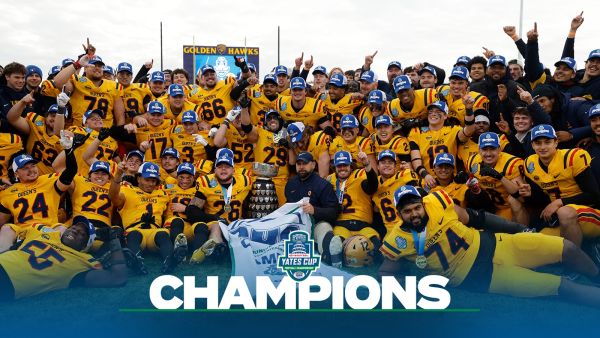 A large university football team in yellow uniforms celebrates together on the field with the Yates Cup trophy, posing under a banner that reads ‘Champions.’