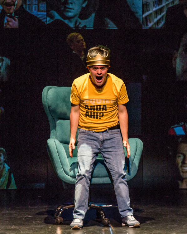 An actor dressed in a yellow shirt and jeans faced towards the camera with arms towards the floor.  