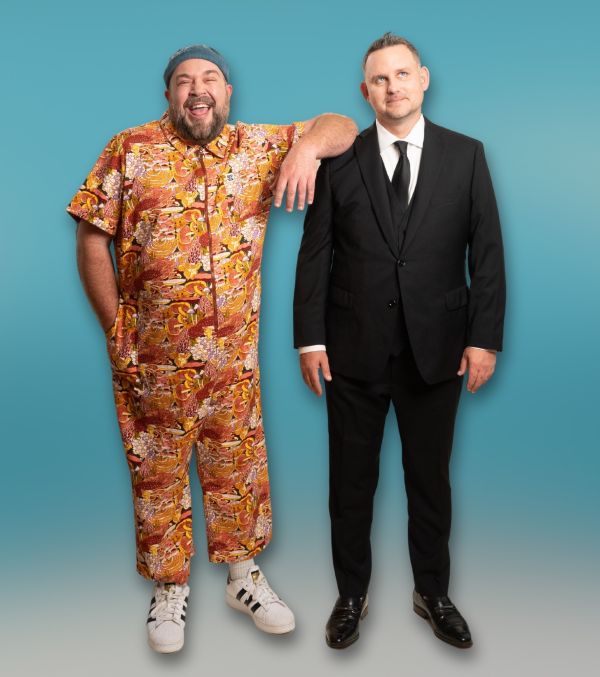Two performers posing together against a blue background, one in a colorful patterned outfit and one in a black suit.