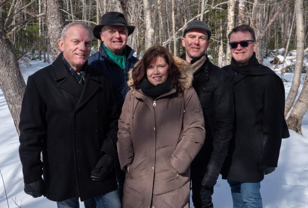 Group of five people standing in a snowy forest, dressed in winter coats and scarves. The leafless trees and snow-covered ground suggest a bright, cold day.