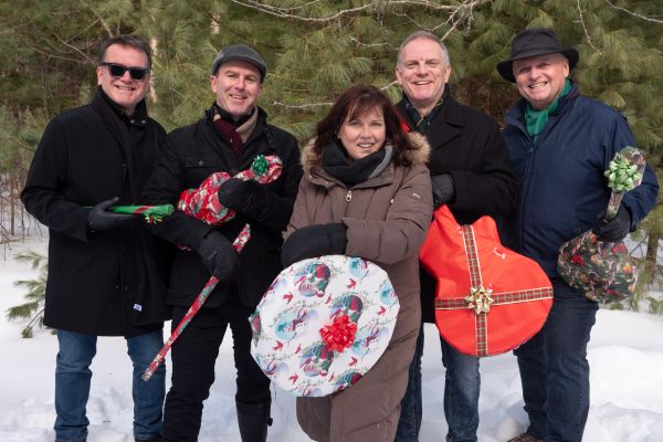 Five people standing in a snowy forest holding wrapped holiday gifts. They are dressed in winter clothing, surrounded by pine trees and festive wrapping, suggesting a seasonal celebration.