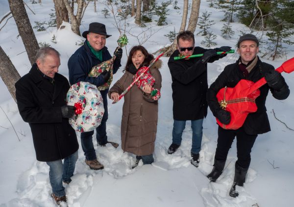 Five people standing in a snowy forest, each holding a musical instrument wrapped in festive holiday paper. They are dressed in winter clothing, surrounded by snow-covered trees, suggesting a seasonal music-themed celebration.