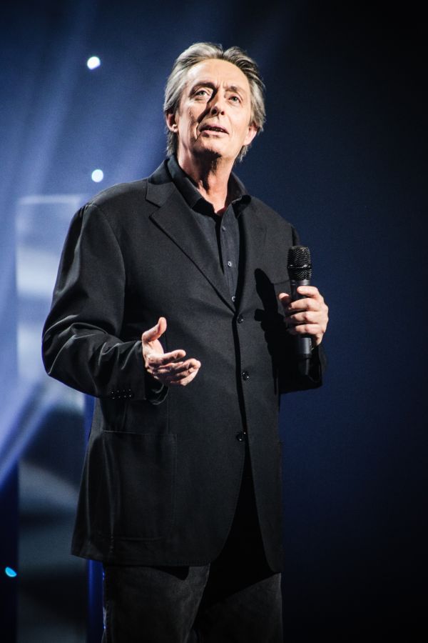 Performer in a dark suit holding a microphone on stage under blue lighting.