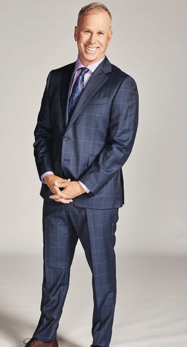 Person (Gerry Dee) wearing a dark blue checkered suit with a light pink shirt and patterned tie, standing against a plain background.