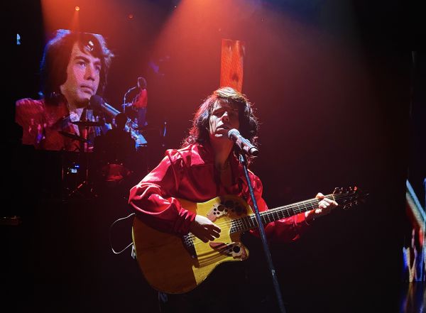 Performer in a red shirt plays an acoustic guitar and sings into a microphone under stage lights, with a large screen projection in the background.