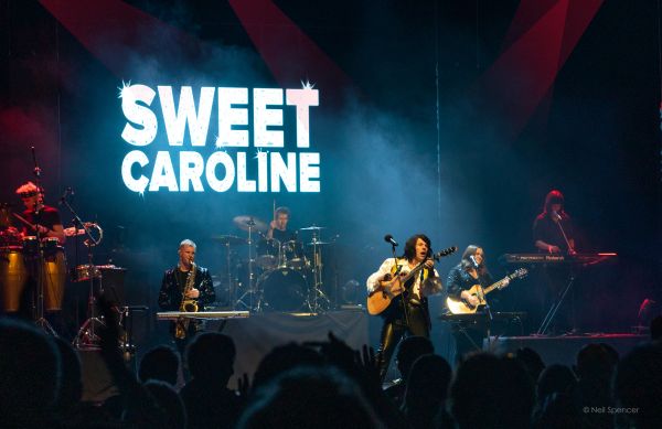 Band performs onstage before a crowd, with “SWEET CAROLINE” displayed on a large screen behind them.