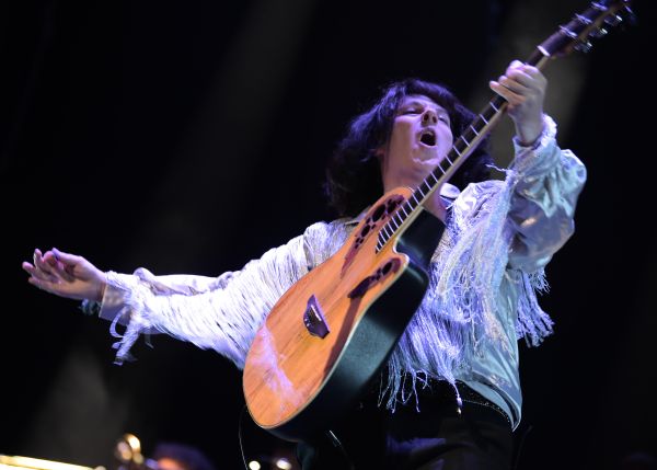 Performer in a fringed white jacket playing an acoustic guitar on a dimly lit stage under spotlights.