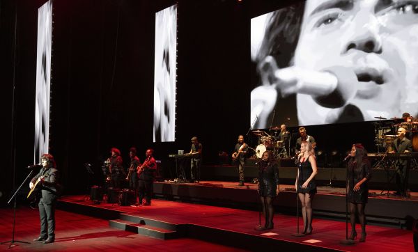 Wide stage performance featuring a guitarist, backing singers, and band with large black-and-white screens in the background.
