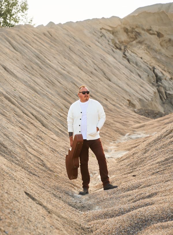 William Prince carrying his guitar case, in a desert gully dressed in brown pants, beige shirt and sweater wearing sunglasses