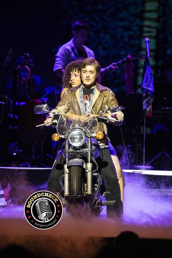 Two performers sit on a motorcycle amid stage fog during a live rock musical performance.