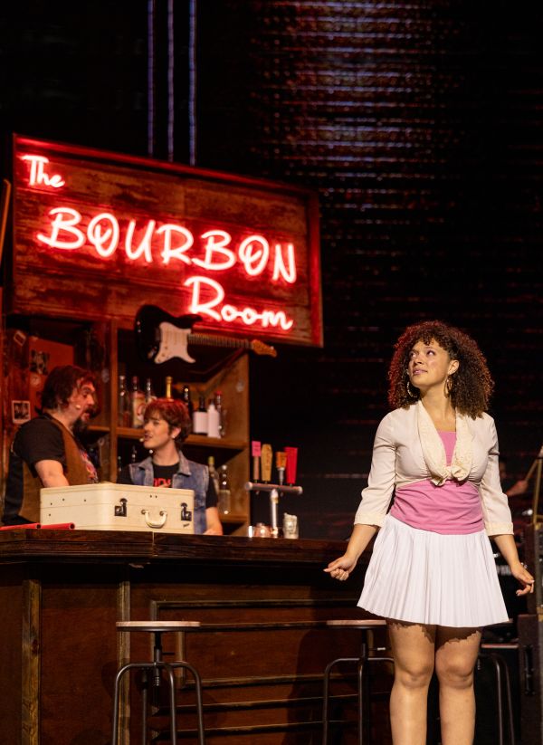 Performer stands onstage beside a bar set beneath a glowing “Bourbon Room” sign during a musical performance.