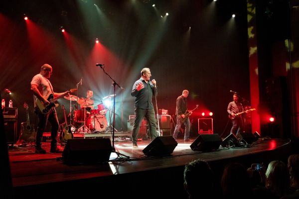 Band performing live on stage under red and white spotlights, with musicians playing guitars and drums and a vocalist at center stage.