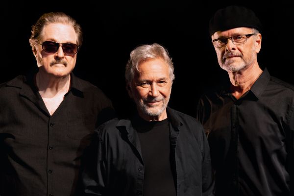 Three performers in black clothing pose together against a dark background.