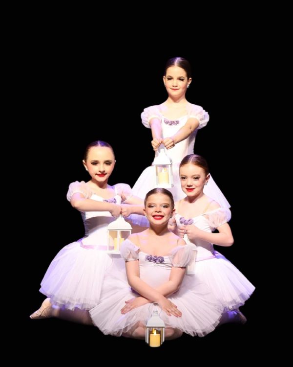 Four young ballet dancers in white tutus pose on a dark stage, each holding a small lantern.