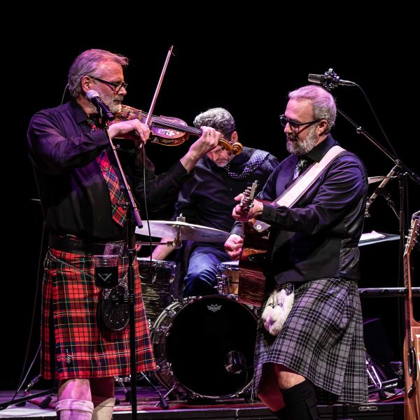 Person playing bagpipes and another with a fiddle on stage with additional musical equipment in the background.